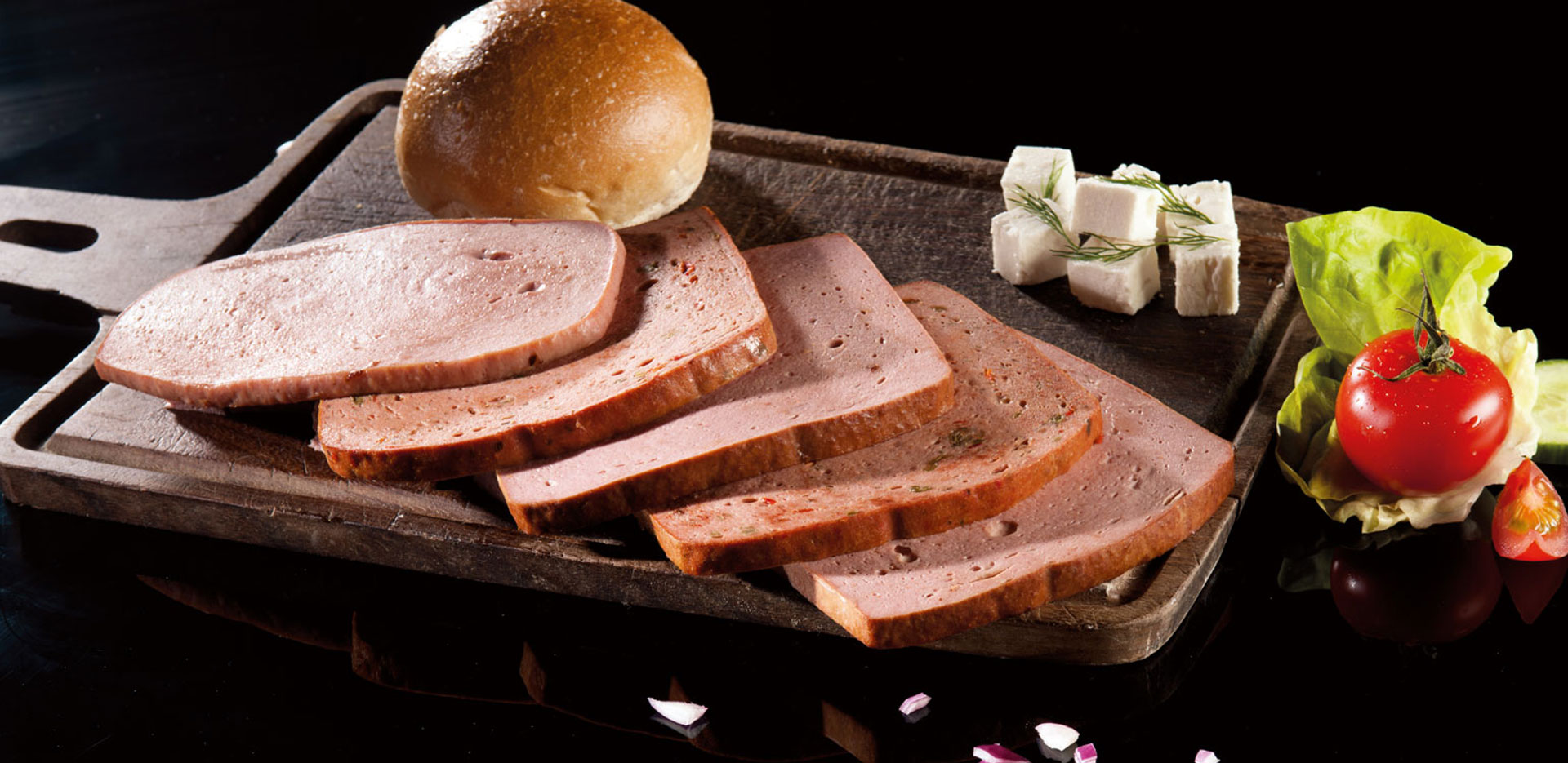 Aufgeschnittene Fleischkäse-Scheiben auf einem Holzbrett, angerichtet mit Brötchen, Käsewürfeln, Dill, Salat, Tomate und Gurke vor schwarzem Hintergrund.