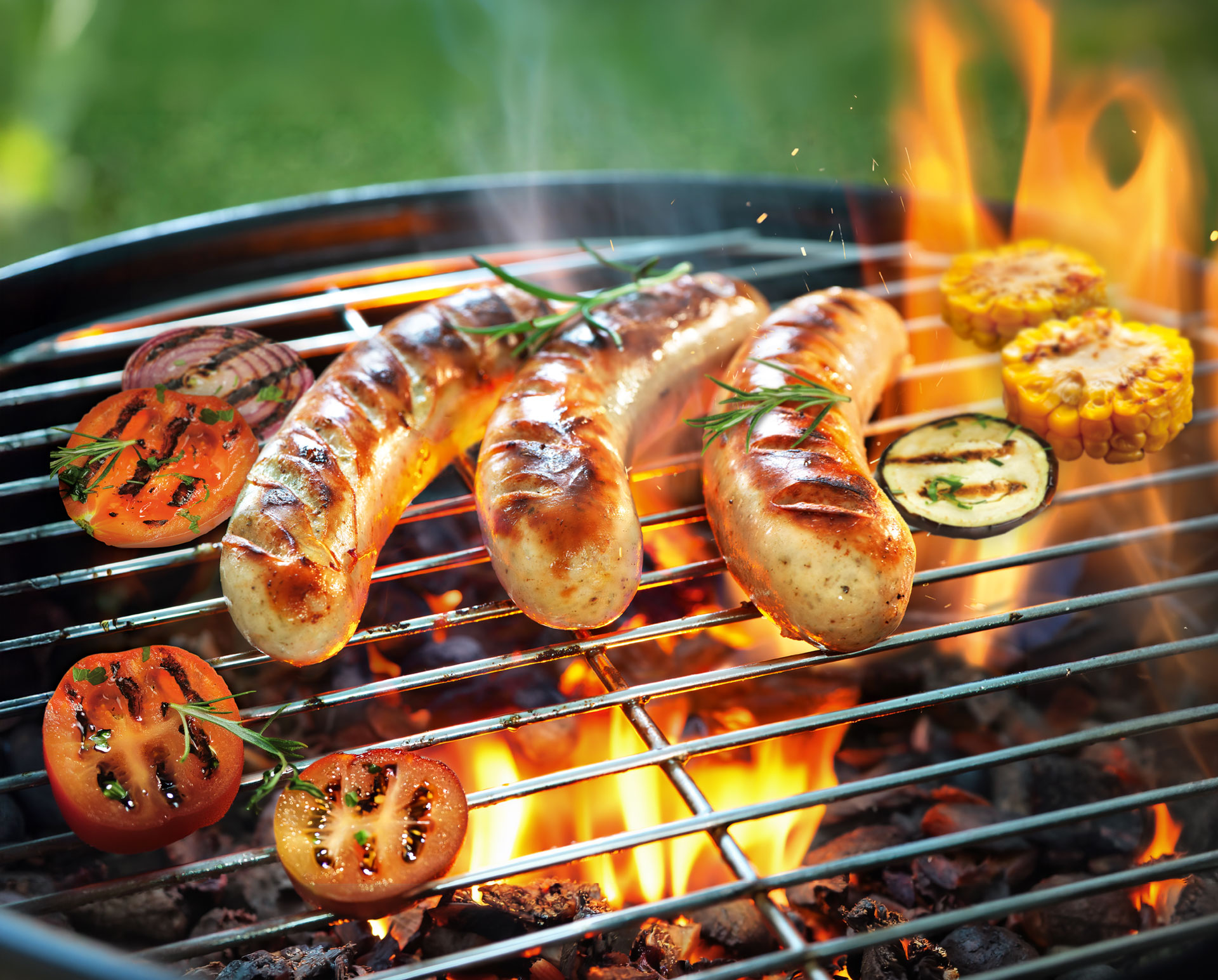Grillrost mit CızzBızz-Bratwürsten, Tomaten, Zwiebel, Aubergine und Maiskolben über offenen Flammen.